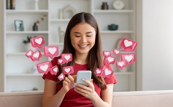 A young chinese woman with long dark hair smiles as she uses her smartphone in a living room setting. Around her are pink hearts with white centers, simulating social media notifications. photo