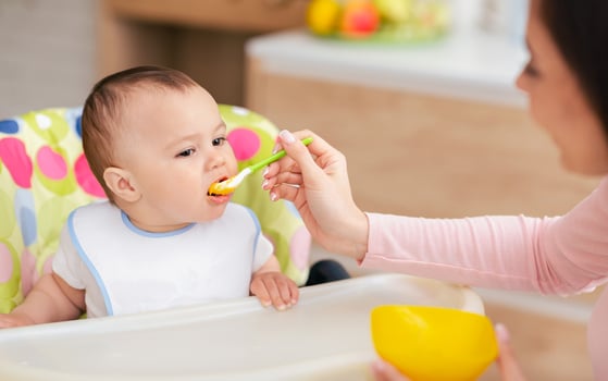 A baby boy is in a child chair eating while his mother feeds him. The kitchen backdrop is cozy and inviting, capturing a tender moment of care and affection. photo