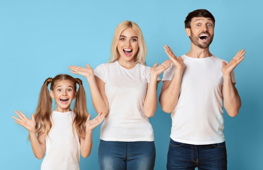 A young girl, a woman, and a man are standing in front of a light blue background. They all have their mouths open in surprise and their arms outstretched, palms up. photo