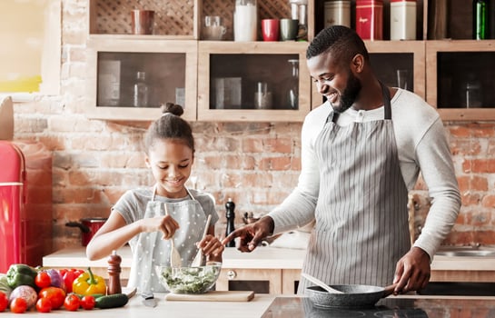 Black smiling girl making veggies salad, dad teaching her, kitchen interior, copy space photo