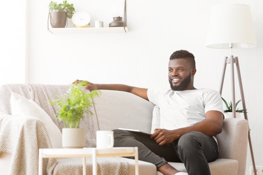 Black man in a white t-shirt and black pants sits relaxed on a cream-colored couch in a brightly lit living room. The room is decorated with a potted plant and a floor lamp photo