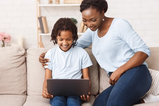 Distance Education Concept. African mother helping her child with homework, using laptop, sitting on sofa at home. Free space photo