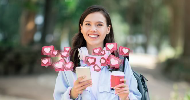 A young woman wearing a light blue shirt and a backpack stands in a park, smiling and looking at her smartphone. She is holding a red to-go cup in her other hand. Surrounding her are red heart icons photo