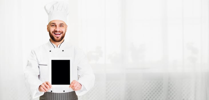 Culinary Recipes App. Chef Cook Man Holding Blank Digital Tablet Screen With Menu Standing In Restaurant Kitchen Indoor. Mockup photo