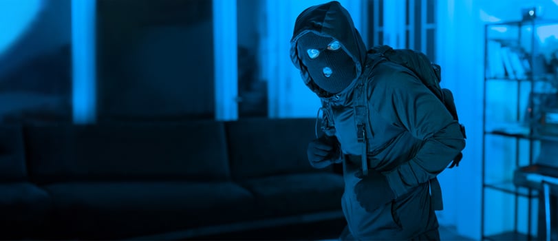 A figure dressed in dark clothing with a hood and mask stands in a dimly lit room at night. The atmosphere is tense, with blue lighting highlighting the scene. photo