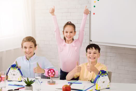 Three young students are sitting at a desk in a classroom setting, giving thumbs up and smiling. The girl in the back is wearing a pink shirt and has her arms raised in the air photo
