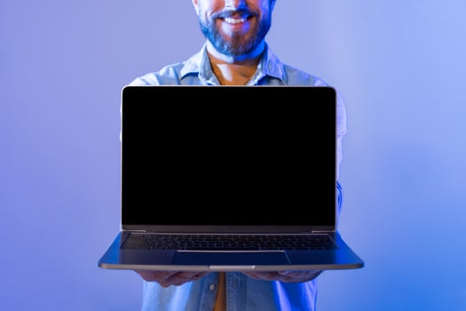 A smiling man with a beard stands against a blue background, holding a laptop with the blank screen facing forward. He appears cheerful and enthusiastic, cropped photo