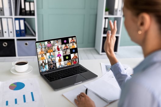 A woman sits at a desk in a home office setting, participating in a conference meeting on her laptop. She has her hand raised as she listens to the other participants on the call photo