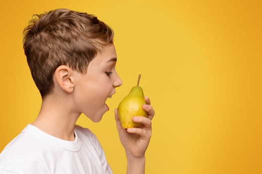 Healthy organic snack. Teen boy biting fresh pear, orange studio background, side view, free space photo