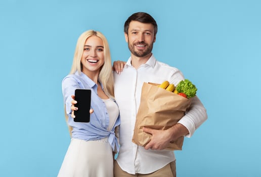 A young woman and a man are standing together against a light blue background. The woman holding a smartphone in front of her with a blank screen, and the man holding a paper bag full of groceries photo