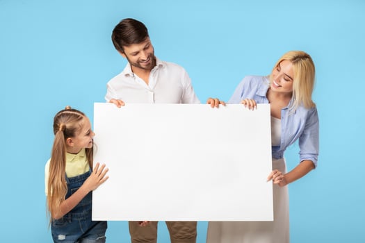 A family consisting of a mother, father, and daughter, are standing in front of a blue background. They are all smiling and holding a large white poster in front of them. photo