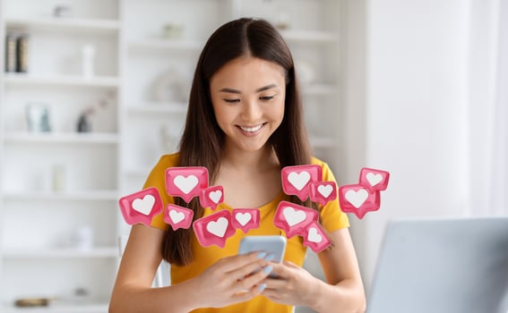 Asian young woman smiles brightly while looking at her smartphone. She is surrounded by animated pink and white heart icons, suggesting that she is receiving many likes on social media photo