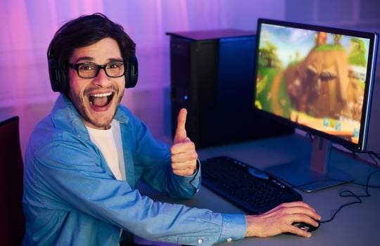 A young man wearing headphones sits in front of a computer, playing a game. He is smiling and giving a thumbs-up gesture, indicating his enjoyment of the game. photo