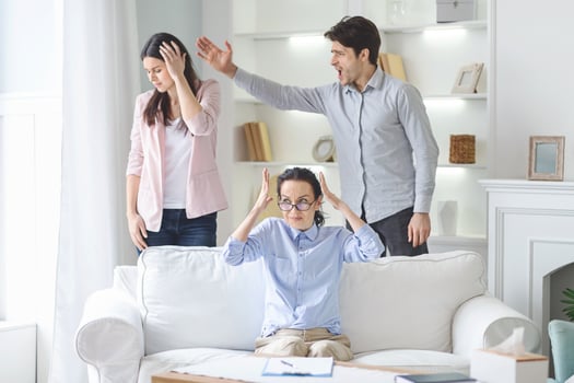 A young couple is seen arguing in a bright, modern living room while their therapist tries to mediate the dispute. The woman covers her face while the man gestures angrily photo