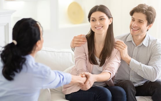 A young couple smiles warmly while having a conversation with a counselor in a cozy, sunlit living room. The counselor is attentively listening and engaging with the couple, shaking hands photo