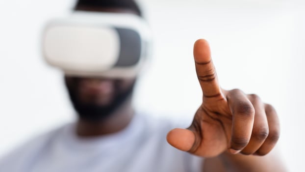 A close-up photo shows African American man wearing a virtual reality headset, his right hand extended forward, with his index finger pointing up as he interacts with digital content.