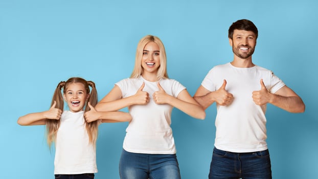 A family of three, a young girl, a woman, and a man, stand in a row in front of a blue background. They all smile brightly and hold their thumbs up in the air. photo