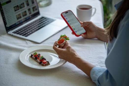 Peaceful woman at table in morning holding sandwich having breakfast with laptop, smartphone. photo
