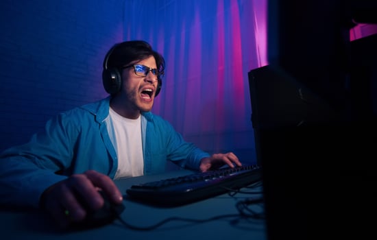 A man wearing headphones and a blue shirt sits in a dimly lit room, intensely focused on playing a game on his computer. The light from the monitor illuminates his face photo