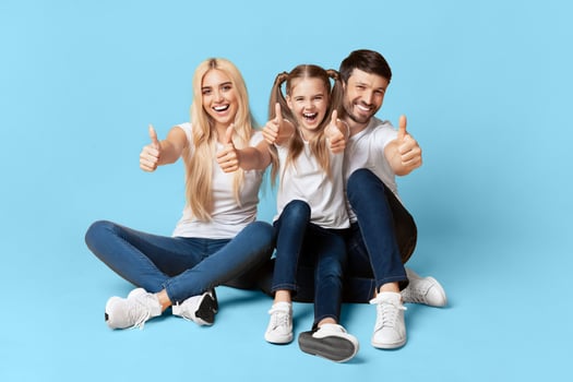 A family of three, a mother, father, and their young daughter, are sitting on a light blue background. They are all smiling and giving the thumbs up. photo
