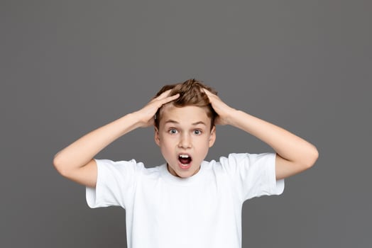 A shocked teen boy is captured with wide eyes and hands clutching his head against an isolated grey backdrop, suggesting alarm photo