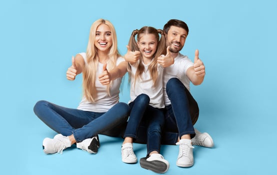 A young family, consisting of a mother, father, and daughter, are sitting on a blue background. They are all smiling and giving thumbs up, conveying a sense of positivity and unity. photo