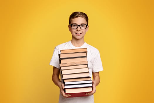 Cute diligent student boy in glasses with heap of books in hands, orange studio background photo
