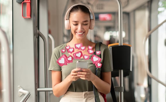 A young woman smiles and looks at her smartphone while wearing headphones. She is standing on a public bus and surrounded by pink heart icons, suggesting she is receiving positive feedback photo