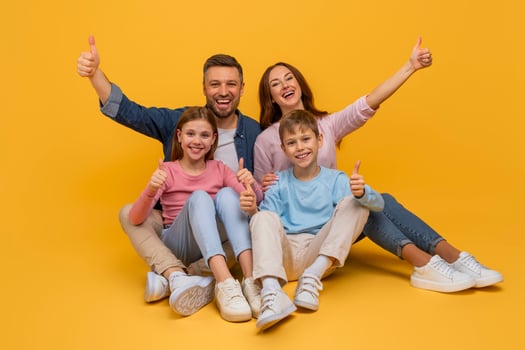A smiling family consisting of a mother, father, daughter, and son are sitting close to each other against a vibrant yellow backdrop, all giving a thumbs-up sign in a show of happiness and unity. photo