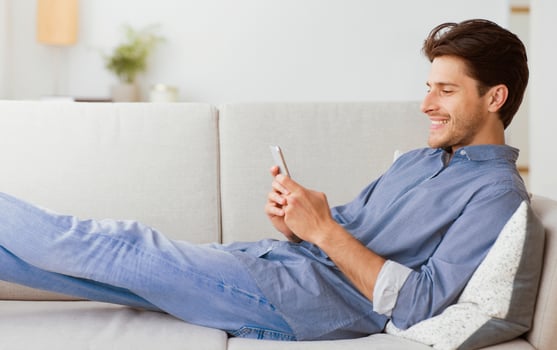 A man in a blue shirt and jeans is relaxing on a white couch while looking at his smartphone. photo