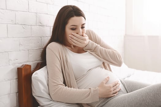 Morning sickness. Young pregnant woman sitting on bed, covering her mouth feeling nauseous during pregnancy, free space photo