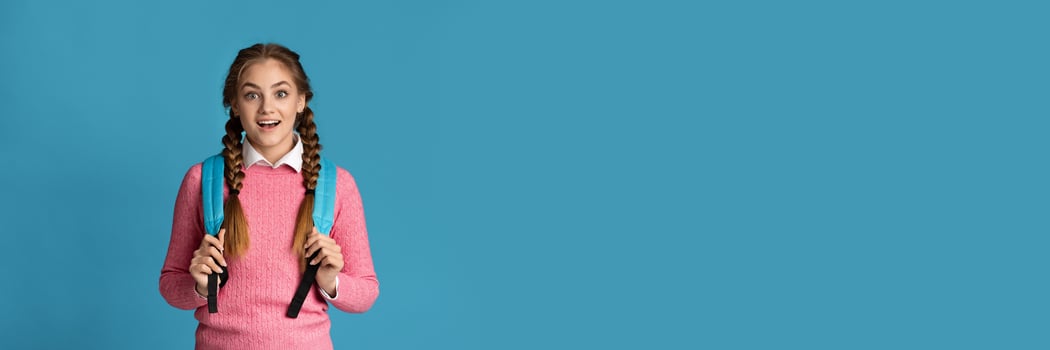 Surprised teenager girl with backpack posing on blue studio background, panorama with copy space. Education concept photo