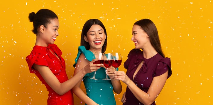 Party Concept. Three Multiracial Women Clinking Red Wine Glasses Laughing And Talking Standing Over Yellow Background. Panorama, Free Space photo