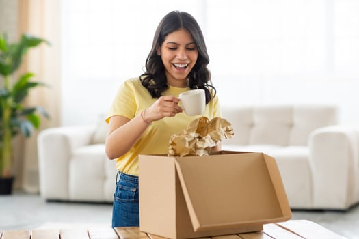 A cheerful young middle eastern woman is standing in a bright, cozy room, enjoying the process of unpacking parcel, holding cup. photo