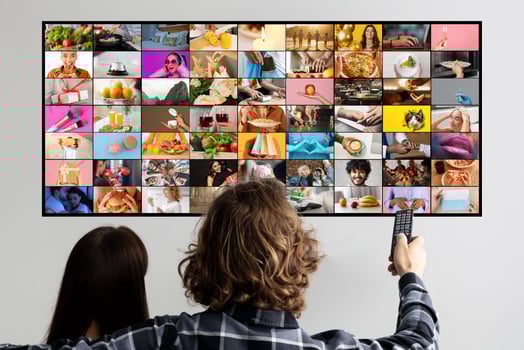 A couple sits on a couch, facing a television screen filled with a collage of images. The woman sits with her back to the camera while the man holds a remote control and faces the screen photo