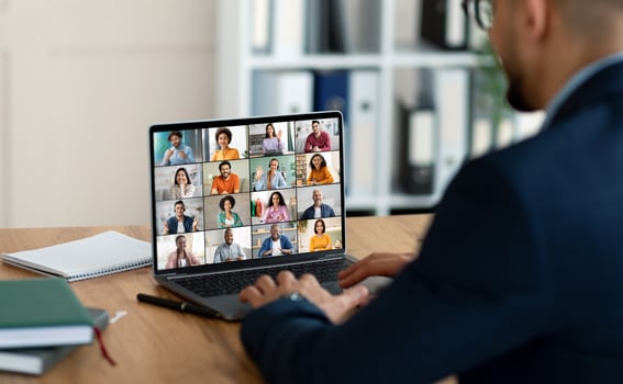 A businessman sits at a desk with a laptop open in front of him, showing a conference screen with many people in individual squares. He is using his laptop for an online meeting. photo