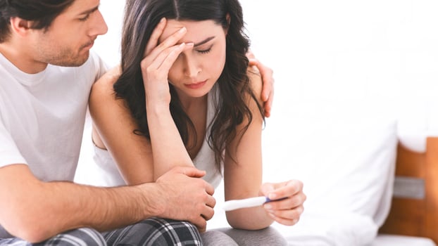 A woman sits on a bed, holding a pregnancy test, with a look of sadness and worry on her face. She is being comforted by her partner, who is placing his arm around her shoulder. photo