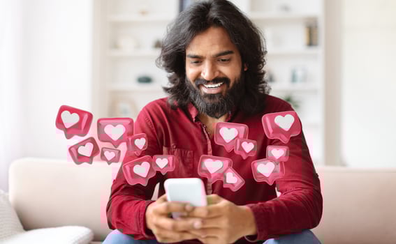 Hindu man with a long beard is sitting on a couch and smiling while looking at his smartphone. There are many heart icons surrounding him, suggesting he is receiving lots of likes photo