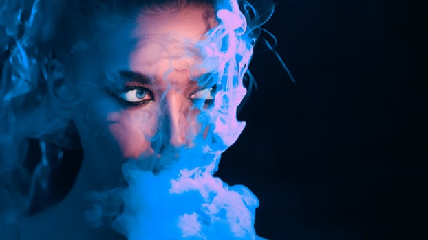 A woman with a captivating, intense gaze peers through swirling blue smoke, creating an air of mystery and intrigue in a dark setting, copy space photo