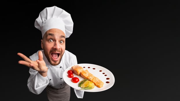 Funny Chef Man Showing Salmon Dish On Plate Posing In Studio On Black Background. High-Angle Shot photo
