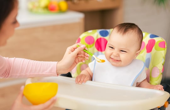 A baby boy is sitting in a high chair being fed by his mother in the kitchen. The atmosphere is warm and comforting, highlighting a loving and nurturing mealtime. photo