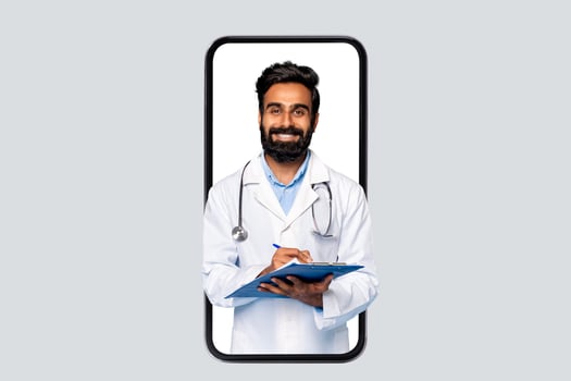 Indian man doctor is seen on the screen of a smartphone, wearing a white coat and stethoscope, smiling, and writing on a blue clipboard with a pen. photo