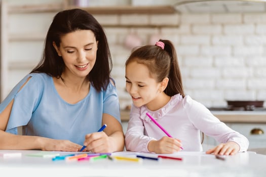 A mother and daughter sit at a table in a kitchen, both concentrating intently on their coloring projects. They are using colorful markers and crayons, and seem to be having fun and bonding photo