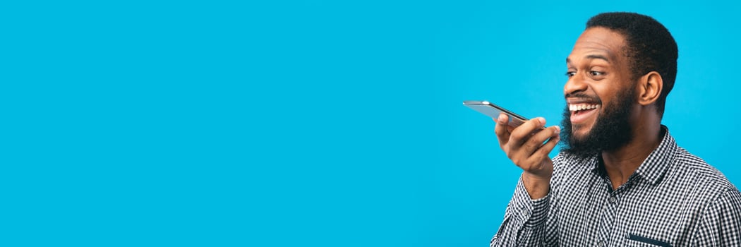 Happy young black man using voice assistant on smartphone, isolated over blue studio wall, copy space, web-banner photo