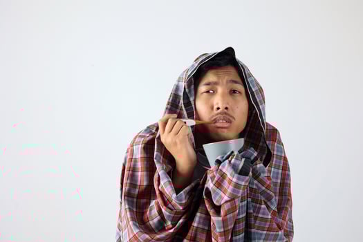 Ill Man Covered with Sarung Holding a Tea Cup and Thermometer photo