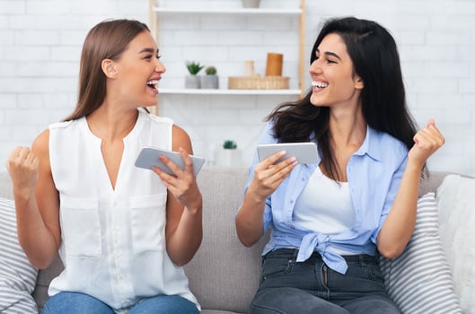Modern Friendship. Two Girls Playing Mobile Games On Phones Gesturing Yes Celebrating Victory Sitting On Couch Indoor. photo