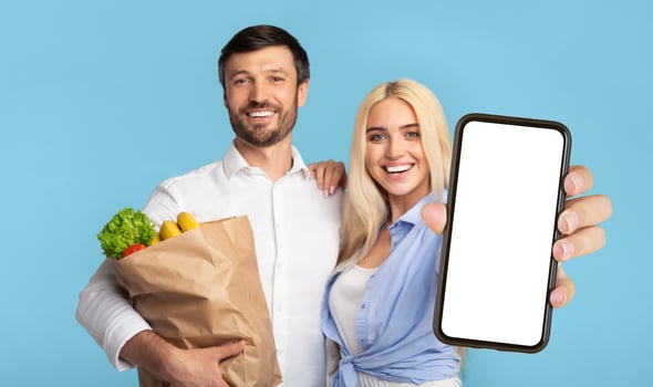 A young man and woman are standing in front of a light blue background. The man is holding a brown paper bag filled with groceries, while the woman is holding a smartphone with a blank white screen photo