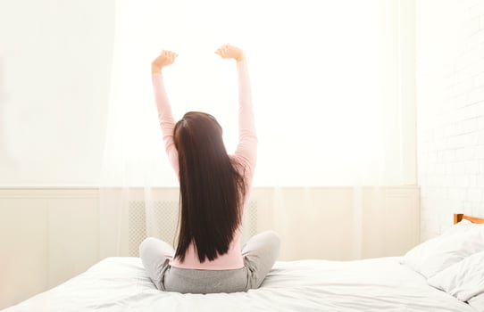 A woman with long dark hair is sitting cross-legged on a white bed, stretching her arms upwards toward the morning sunlight streaming through a large window, back view photo