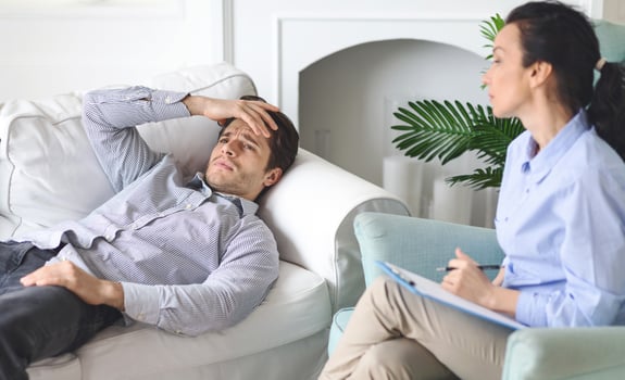 Frustrated man is seen discussing his thoughts with a therapist in a cozy living room. He appears distressed, resting his head on his hand, while the therapist listens attentively and takes notes. photo