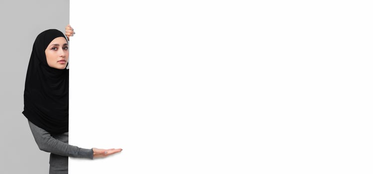 Serious arabic girl in headscarf pointing at white blank advertisement board with open palm, grey studio background photo
